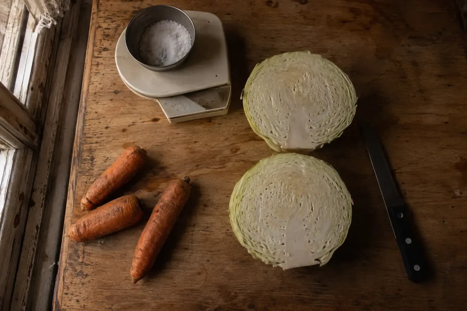 Кочан белокочанной капусты, разрезанный пополам, рядом кухонные весы с солью, три морковки и нож